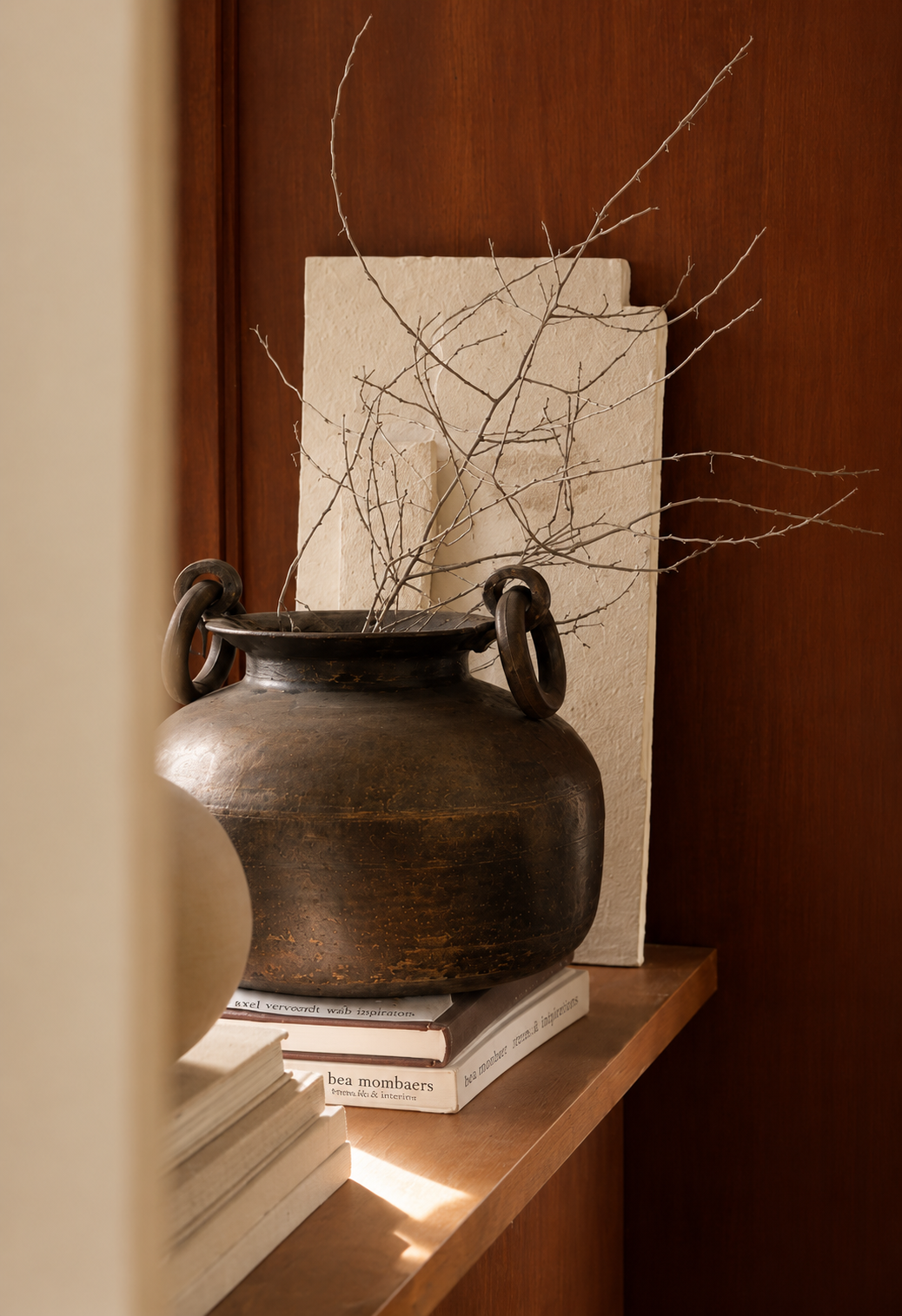 Quiet wabi-sabi vignette: aged brass pot, stacked design books and dried branches on a wooden shelf